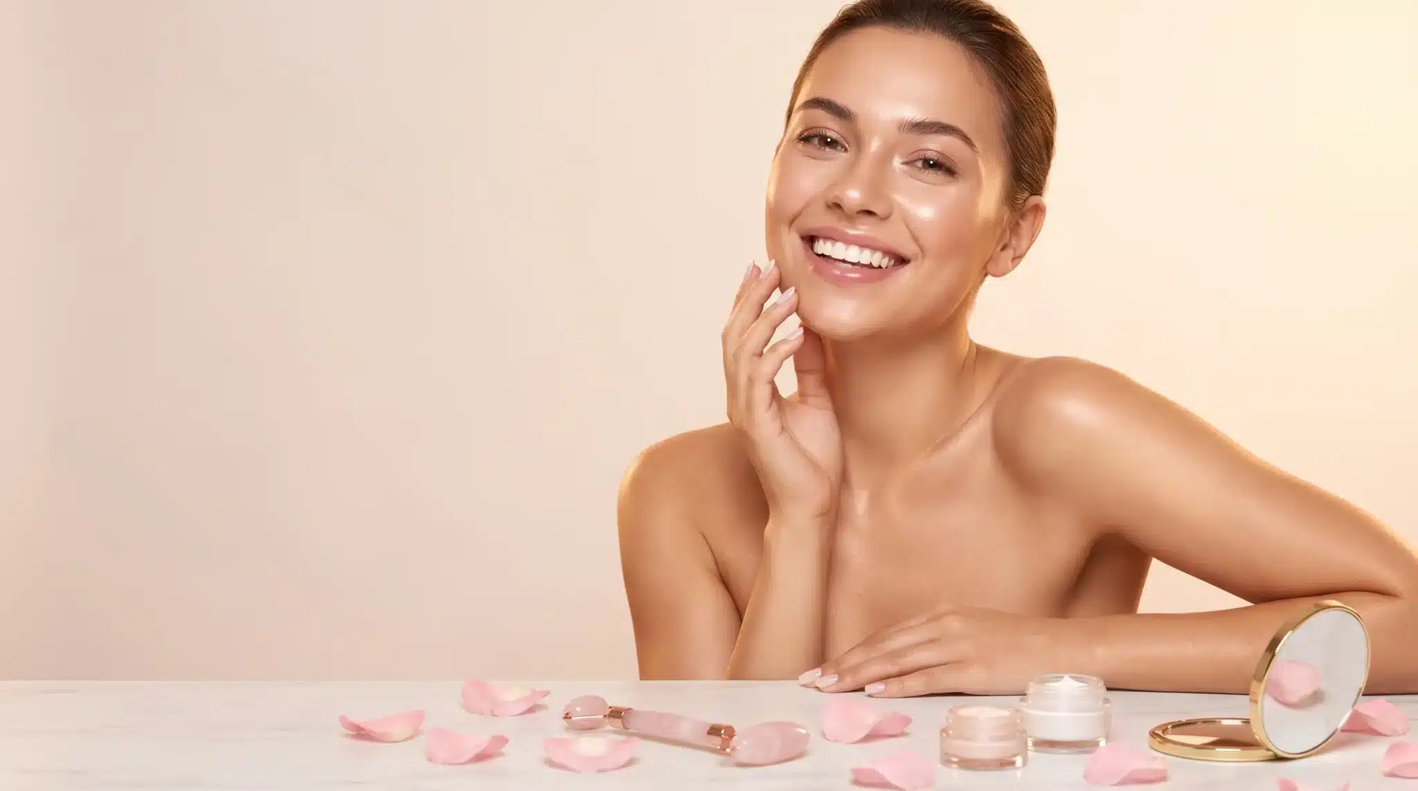 Woman with natural makeup smiling, surrounded by skincare jars and pink rose petals on marble surface