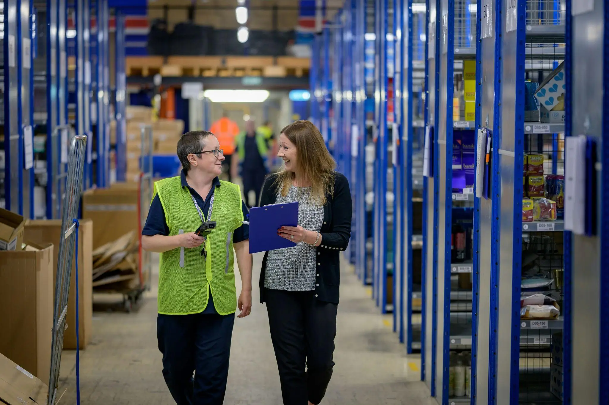 Two colleagues walking and discussing in a brightly lit warehouse aisle with blue shelving