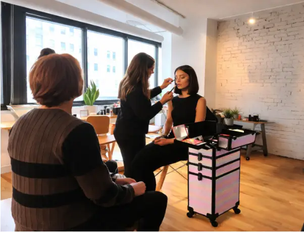Makeup artist applying cosmetics to woman in bright studio with exposed brick wall