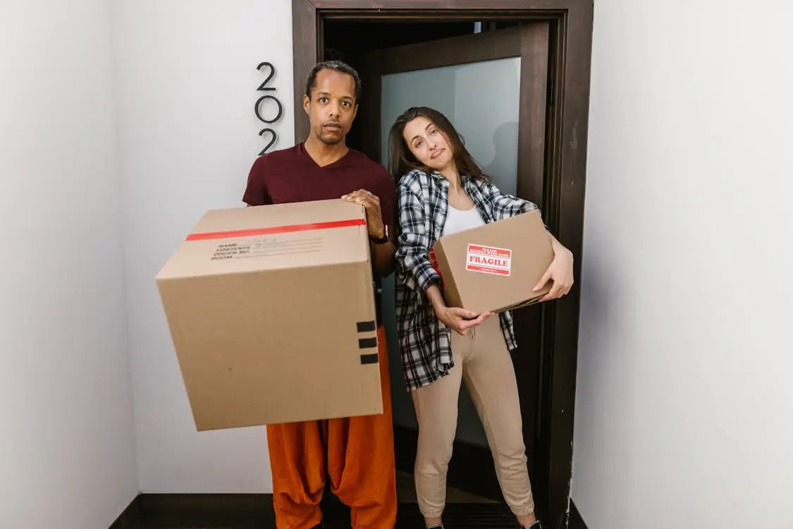 Two people holding cardboard boxes at doorway of apartment 202