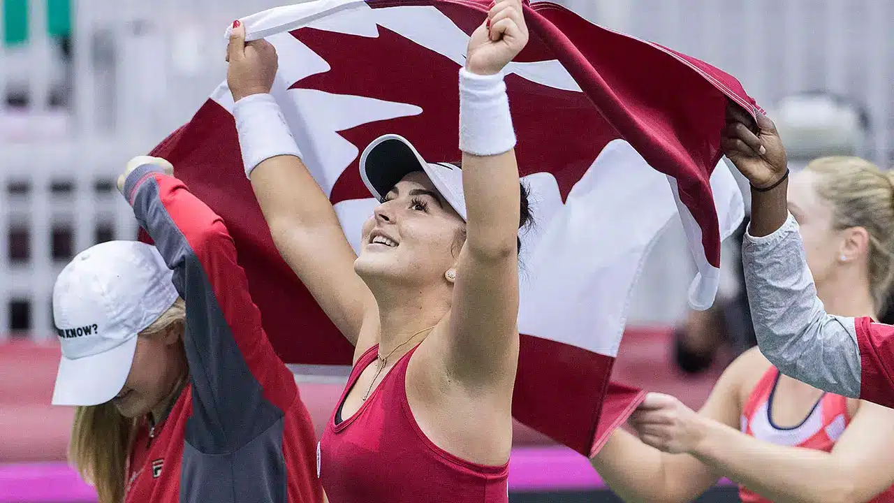 Tennis player holding Canadian flag overhead on indoor court with teammates nearby