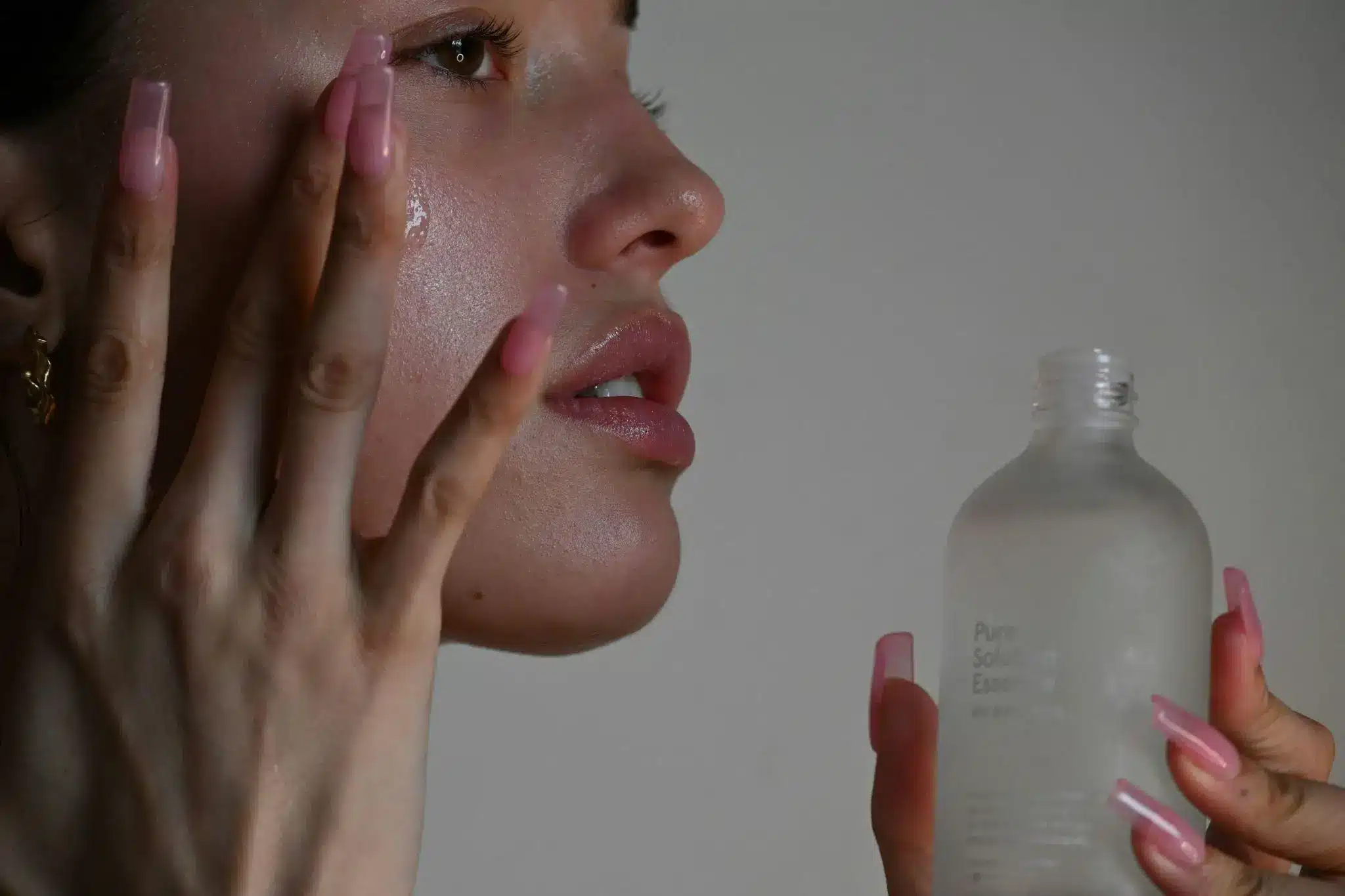 Close-up of person applying facial serum in dim lighting with pink acrylic nails