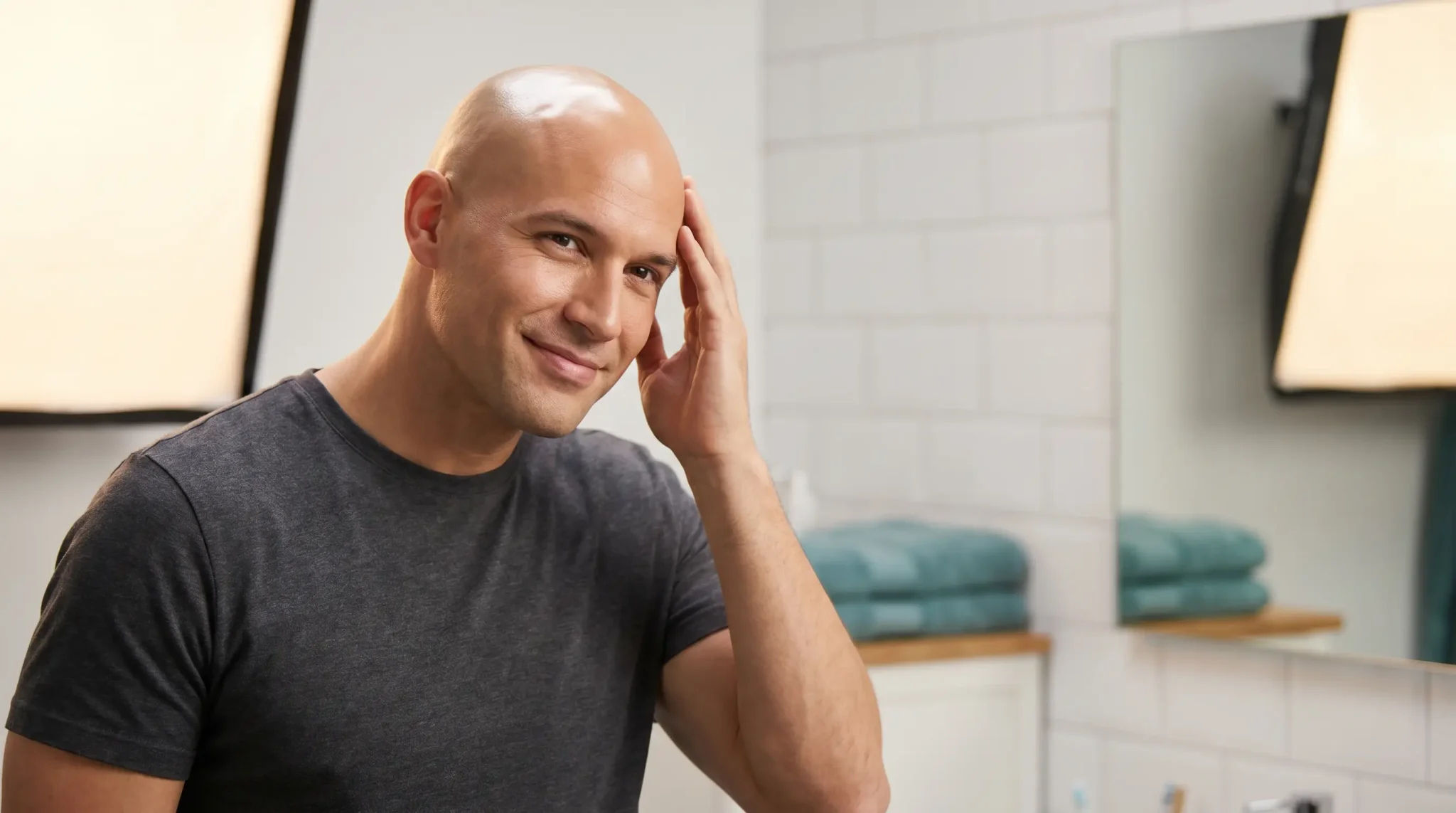 Bald man in gray shirt smiling and touching head in bright bathroom with towel stack
