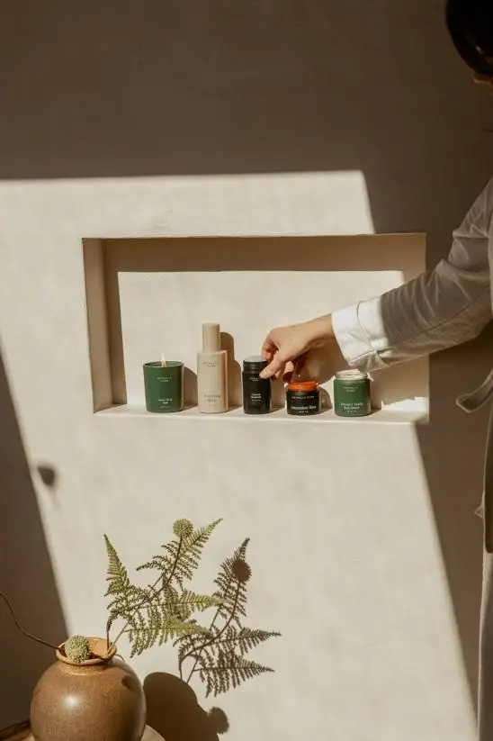 Hand reaching for candles and bottles in minimalist niche with sunlight and fern shadow