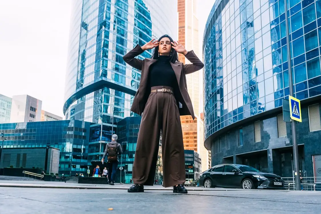 Person in brown suit posing confidently in front of modern glass skyscrapers in urban setting