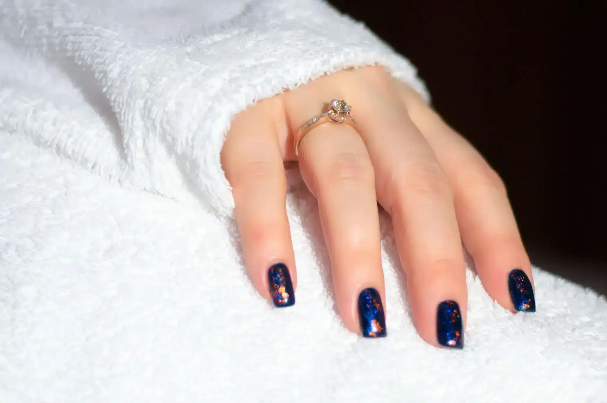 Close-up of a hand with a diamond ring and dark glittery nail polish on white fabric