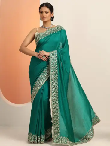 Woman in embroidered teal saree standing indoors against a light backdrop