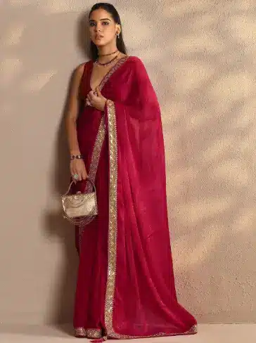 Woman in red sari with gold trim standing against textured beige wall holding handbag