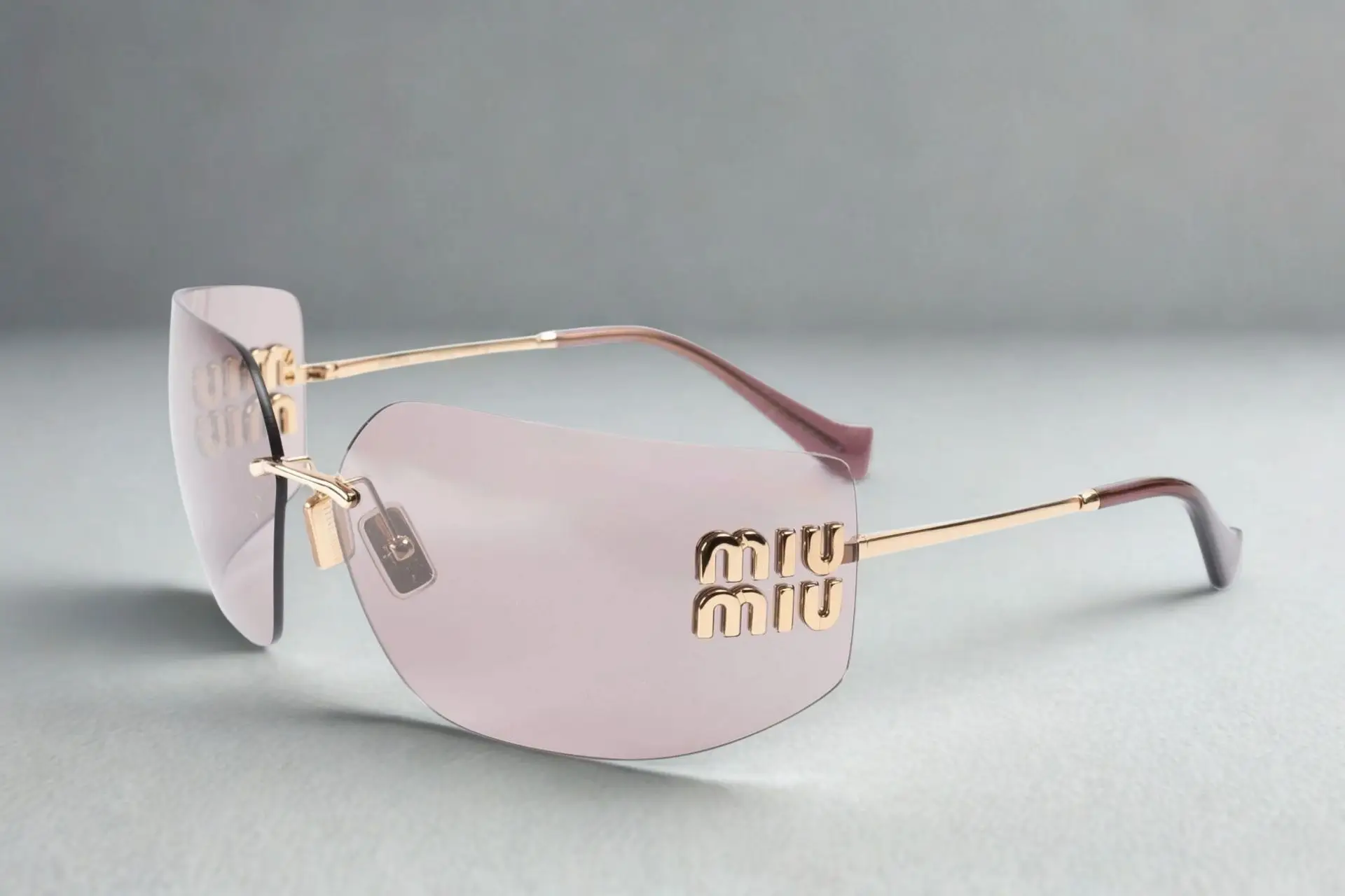 Purple-tinted sunglasses with gold metal frames and logo detail on light surface