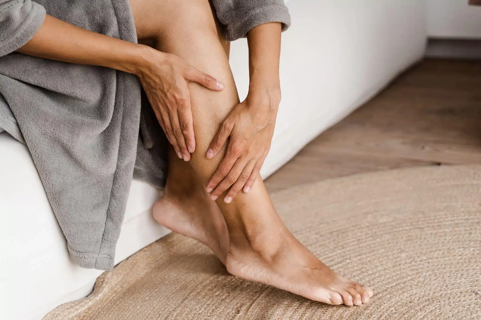 Hands massaging calf of bare leg while sitting on beige rug in cozy room