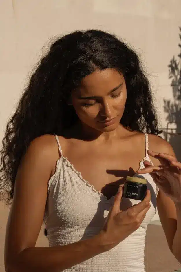 Woman applying balm from jar in warm sunlight with shadowed wall background