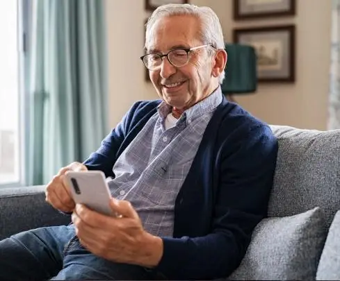 Elderly man smiling while using smartphone on gray sofa in cozy living room