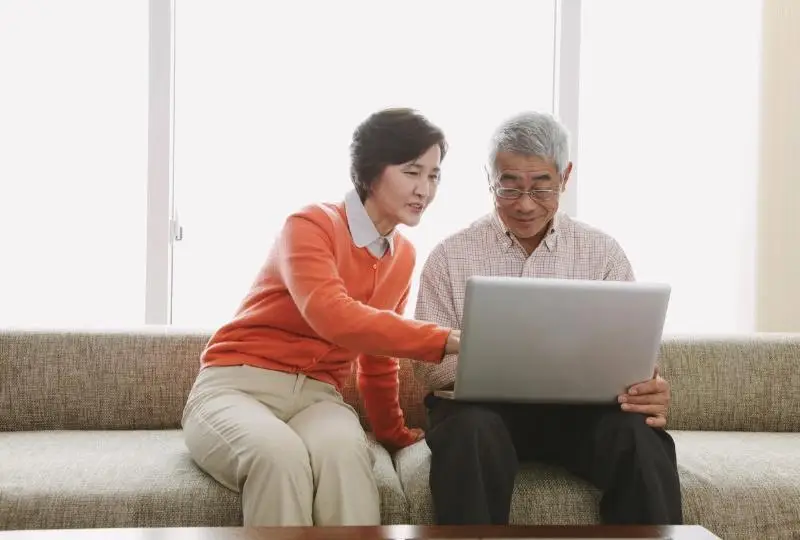 Elderly couple using a laptop together on a beige sofa in a bright living room