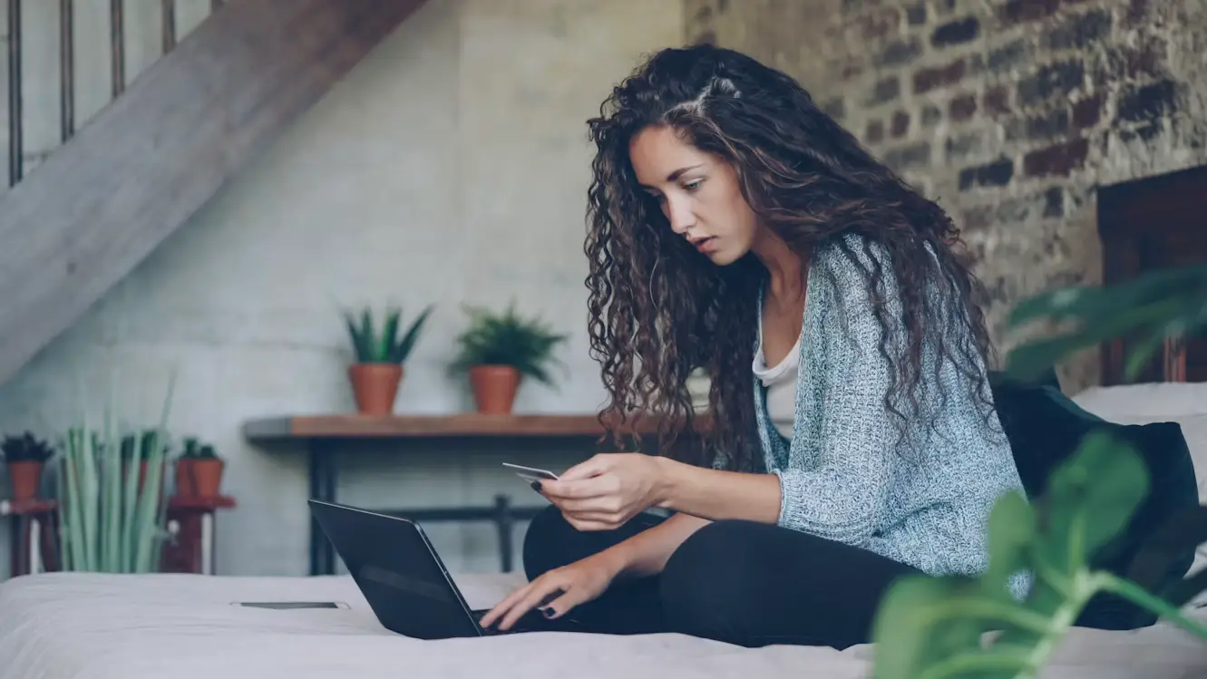 Woman with curly hair shopping online using laptop and credit card in cozy bedroom setting