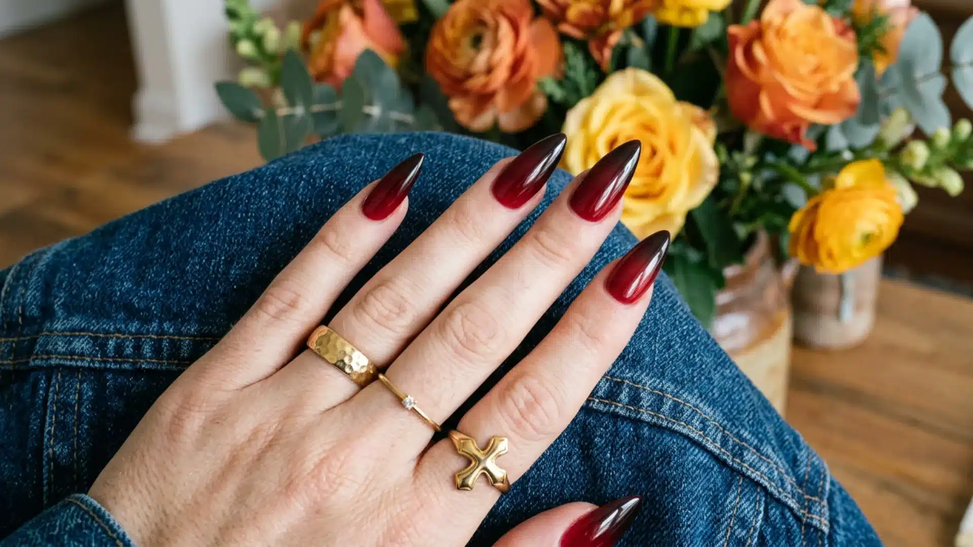 Hand displaying bold wine red jelly nails with sharp pointed shapes and stylish gold jewelry against a floral background