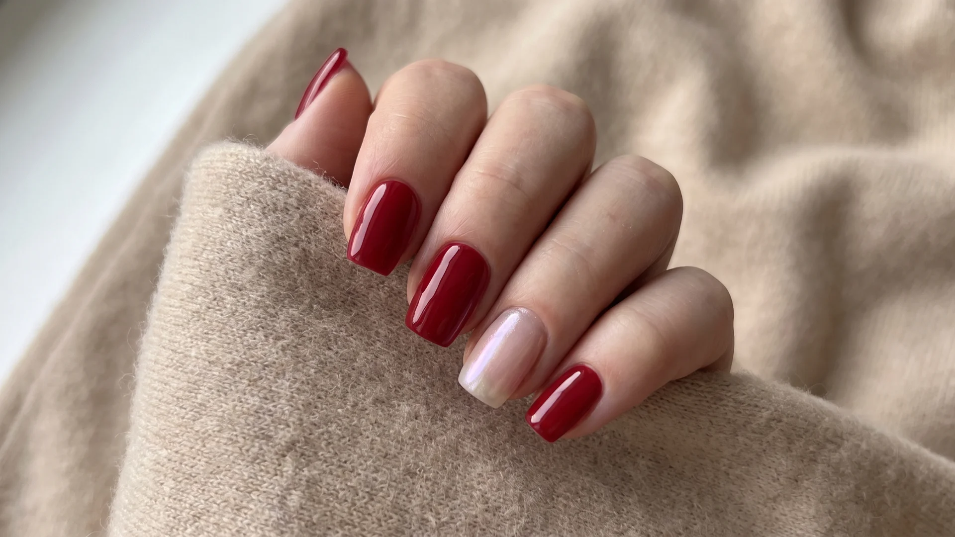 Classic cherry red manicure with one elegant pearly nail accent displayed against a warm knitted fabric background