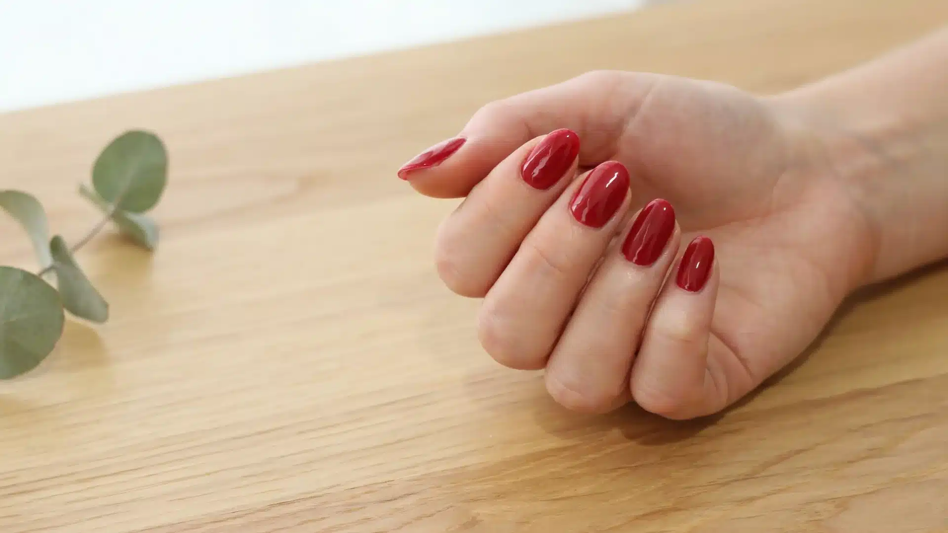 Classic red manicure featuring polished nails on a hand placed over a light wood table with subtle greenery nearby