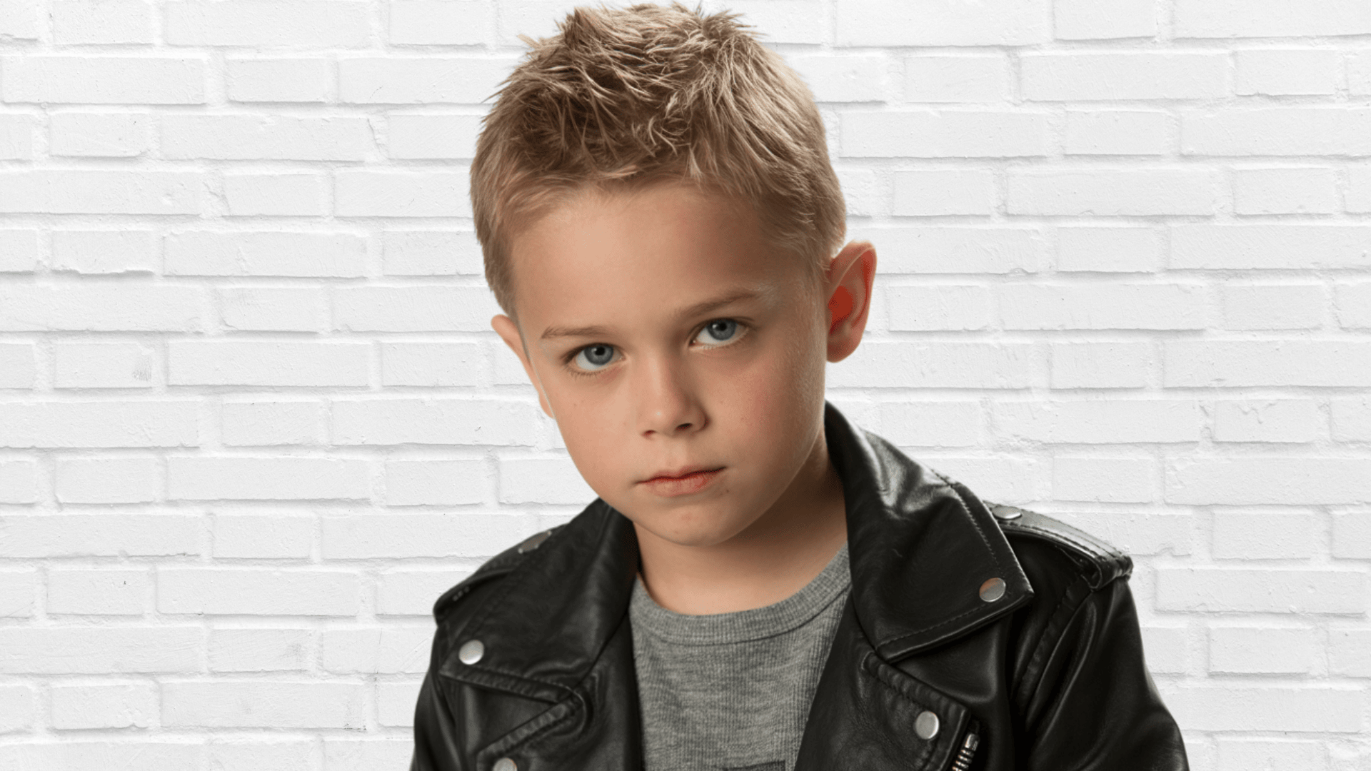 Young boy with crew cut hairstyle wearing a black leather jacket against a white brick wall backdrop.