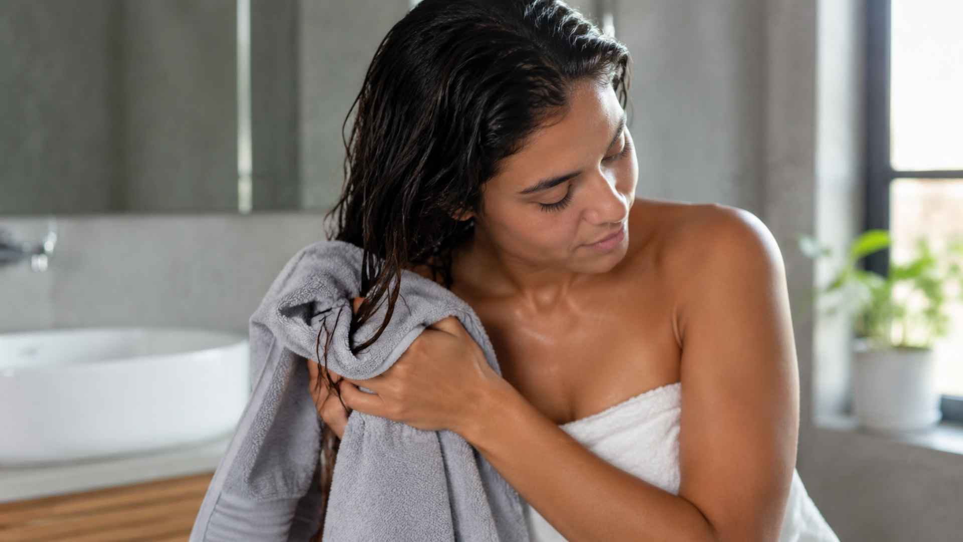 Woman gently drying wet hair with microfiber towel in a bright bathroom after washing hair
