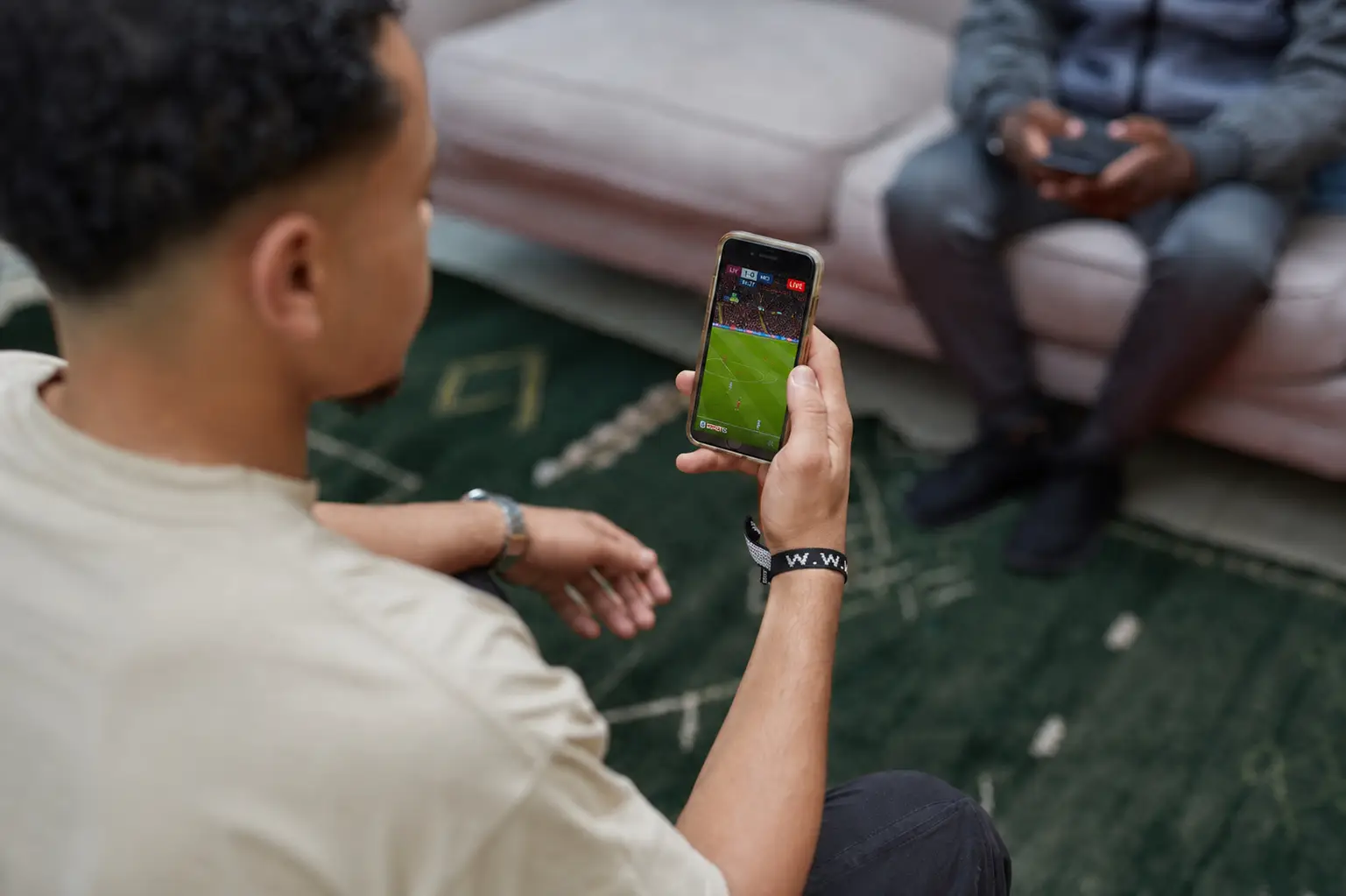 Person watching live soccer match on smartphone in living room with green patterned rug