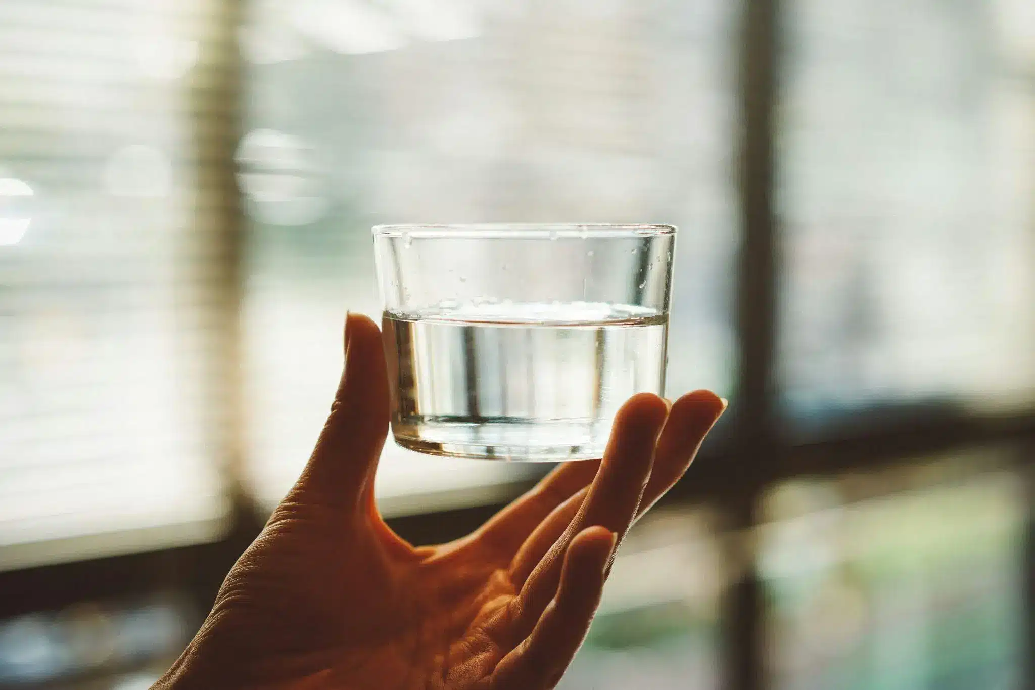 Hand holding glass of water against a blurred window background with natural light