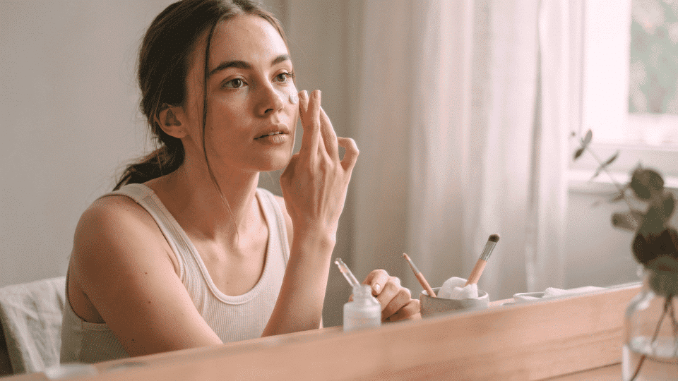 A woman sitting at a vanity while applying skincare and makeup preparation before a glowy makeup routine