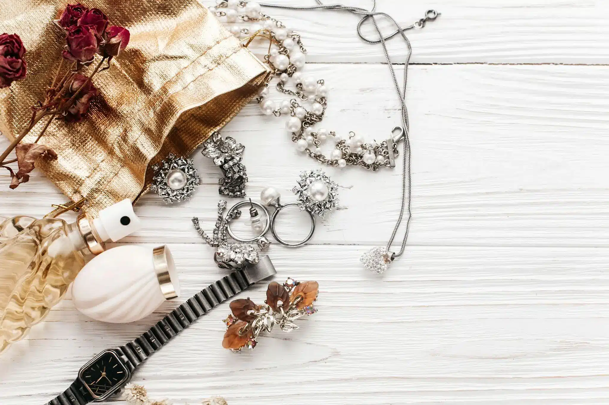 Assorted jewelry and perfume on white wooden surface with gold fabric and dried flowers