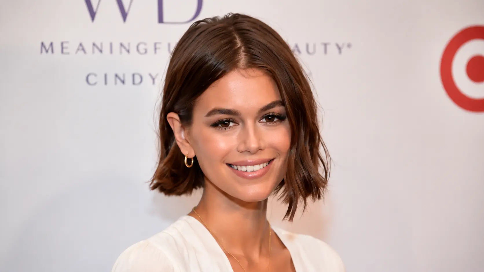 Kaia Gerber with short brown hair wearing a white outfit, posing at an event with a branded backdrop.