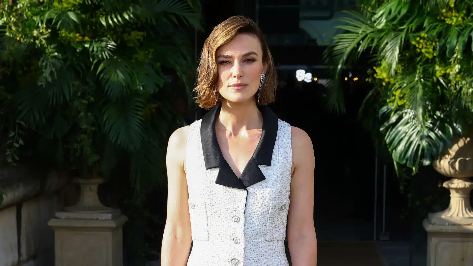 Keira Knightley wearing a white sleeveless dress with black collar detail posing outdoors among green plants at an event