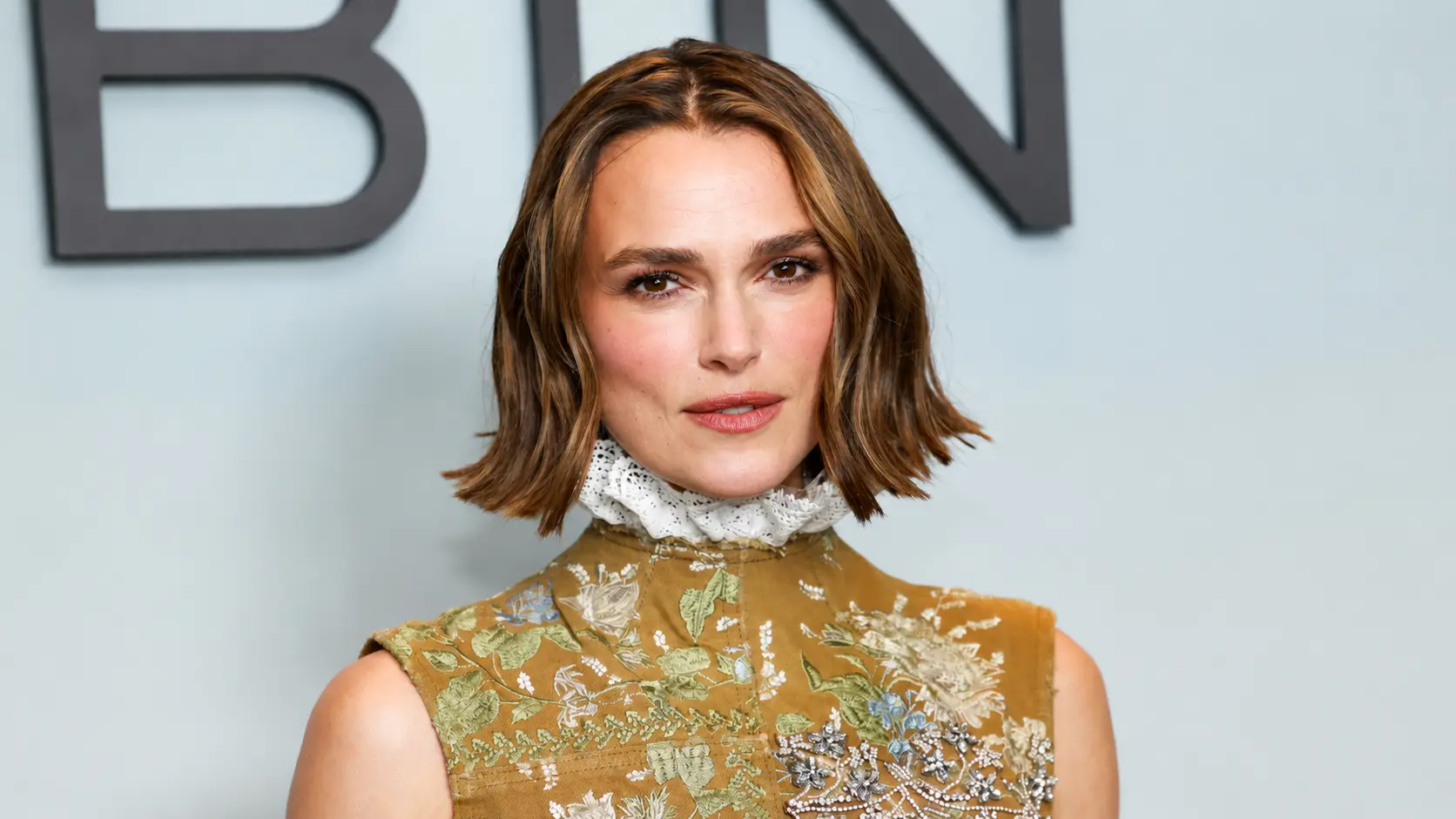 Keira Knightley with shoulder-length brown hair wearing an embroidered gold dress with lace details, posing against a light backdrop.