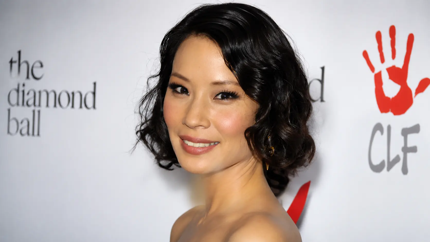 Lucy Liu with short black wavy hair smiling at a red carpet event, wearing earrings against a white branded backdrop.