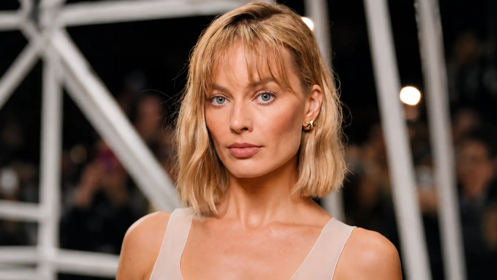 Margot Robbie with bob haircut poses at an event, wearing a sheer top against a dark geometric backdrop.