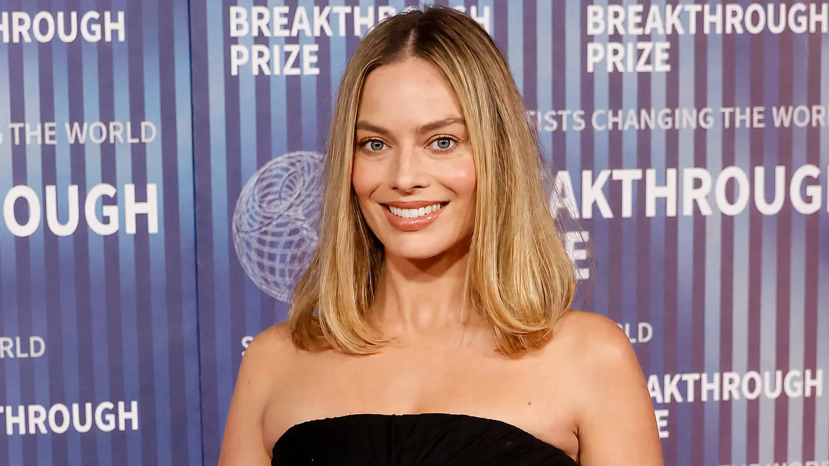 Margot Robbie in a black strapless outfit posing at the Breakthrough Prize event with a branded backdrop.