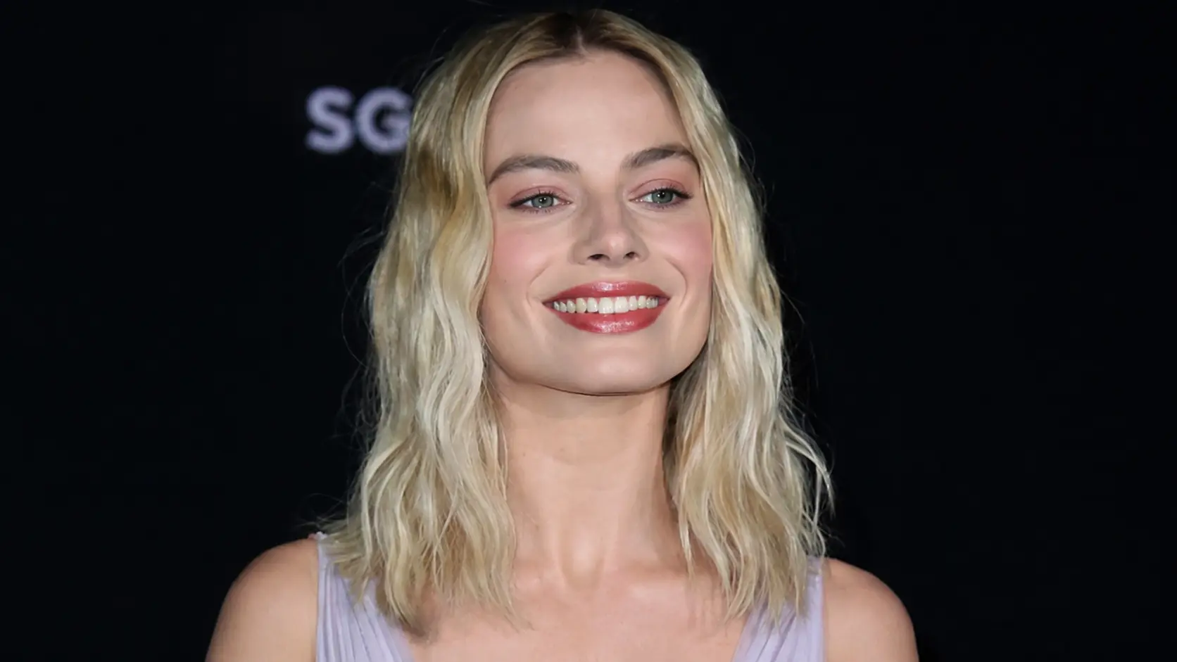 Margot Robbie in a lavender dress posing at an event against a dark background with soft lighting.