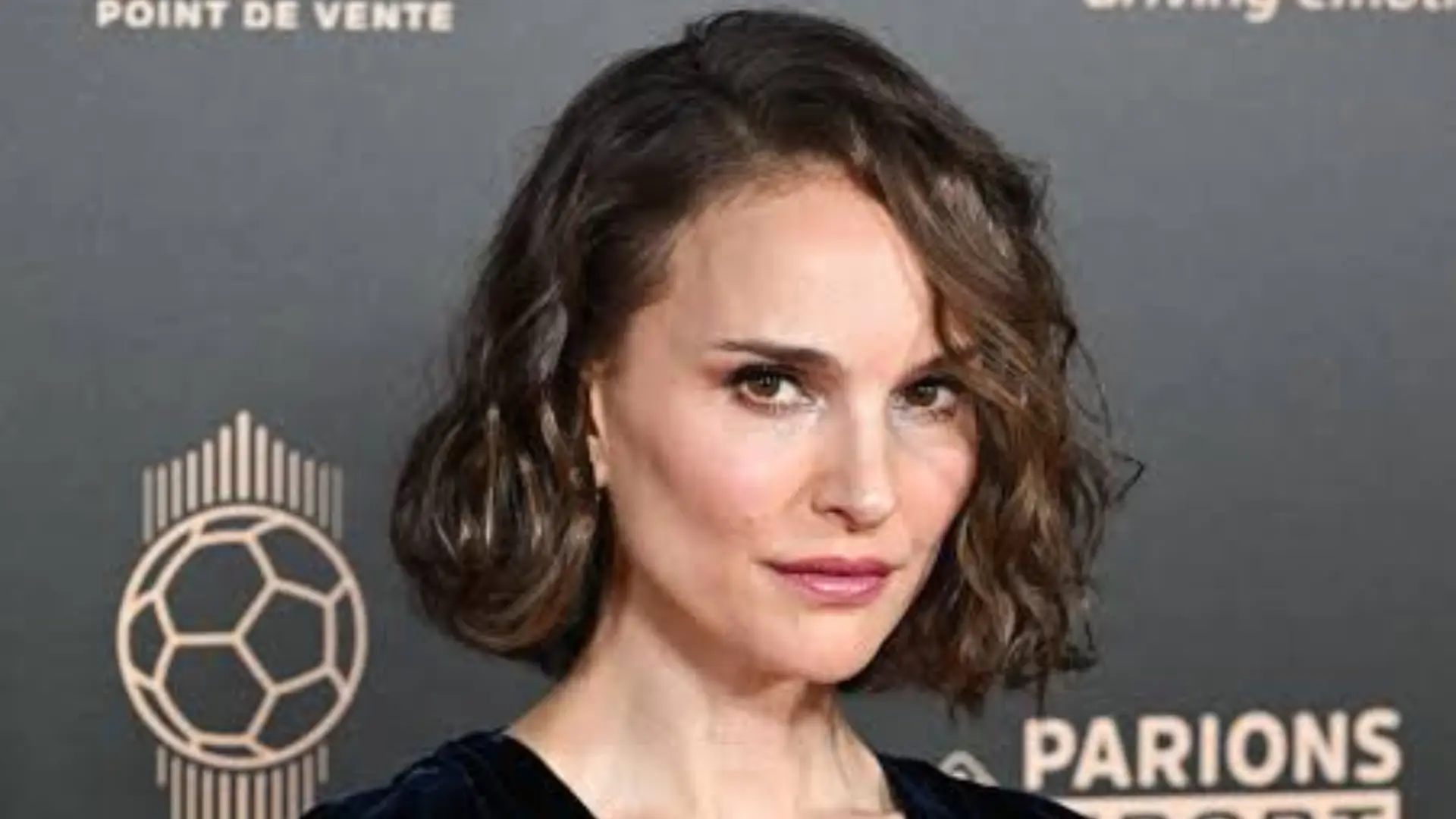 Natalie Portman wearing a black velvet outfit with short wavy hair posing at a sports event backdrop