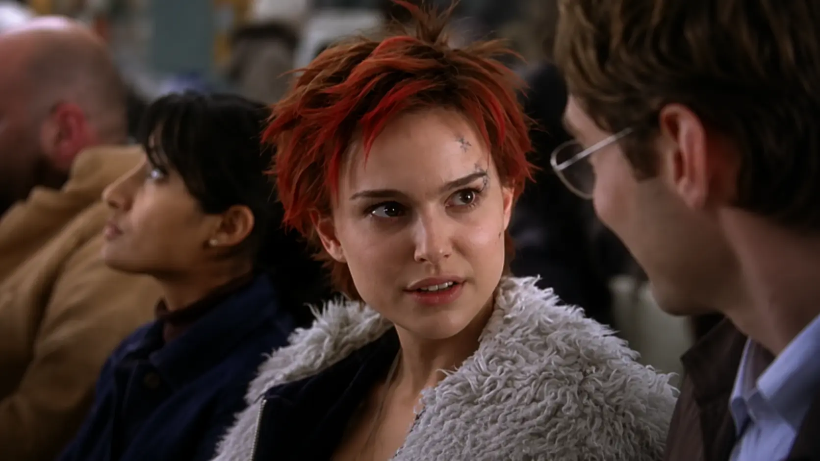 Natalie Portman with short red hair and a white coat sitting in a crowded setting, looking toward another person in a cinematic scene.