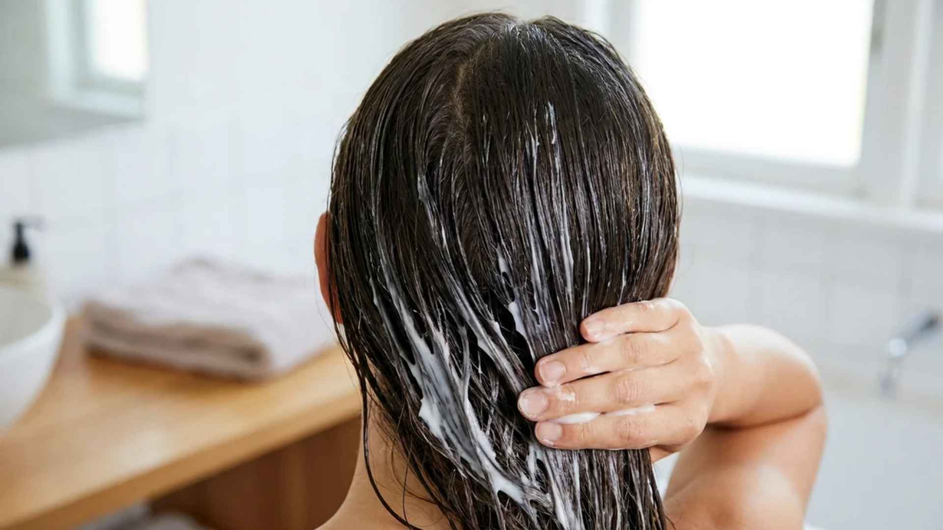 Hands applying conditioner to damp hair lengths for moisture and hydration in a clean bathroom setting