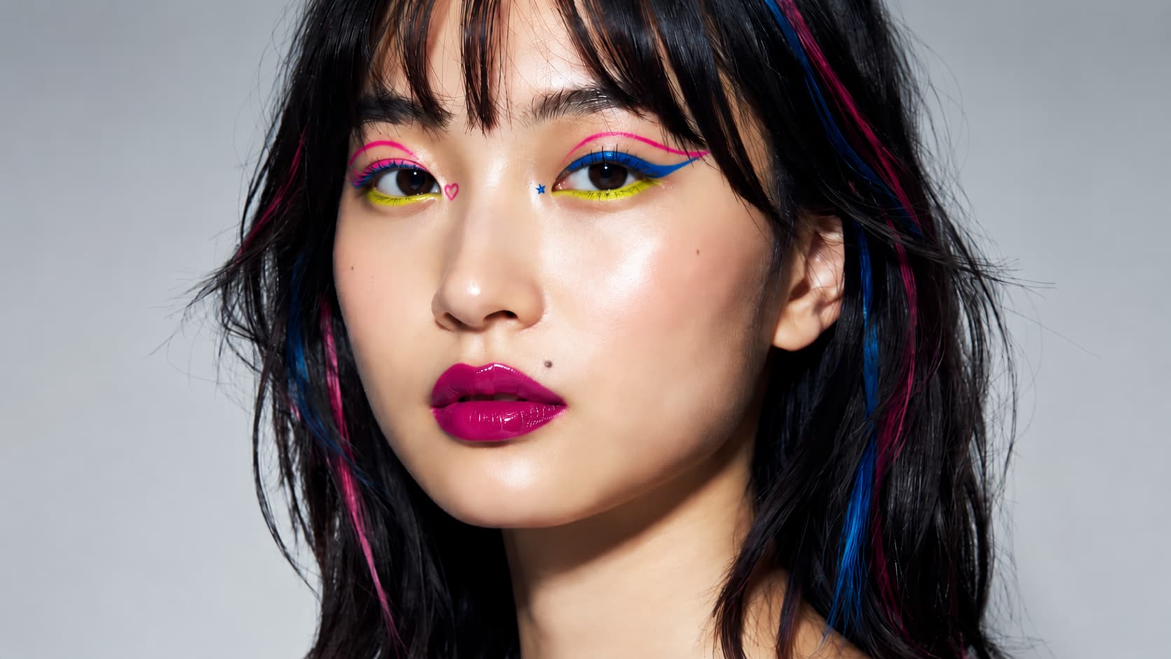 Woman with neon pink and blue winged eyeliner, yellow eyeshadow, glossy fuchsia lips, and pink-blue streaked hair.
