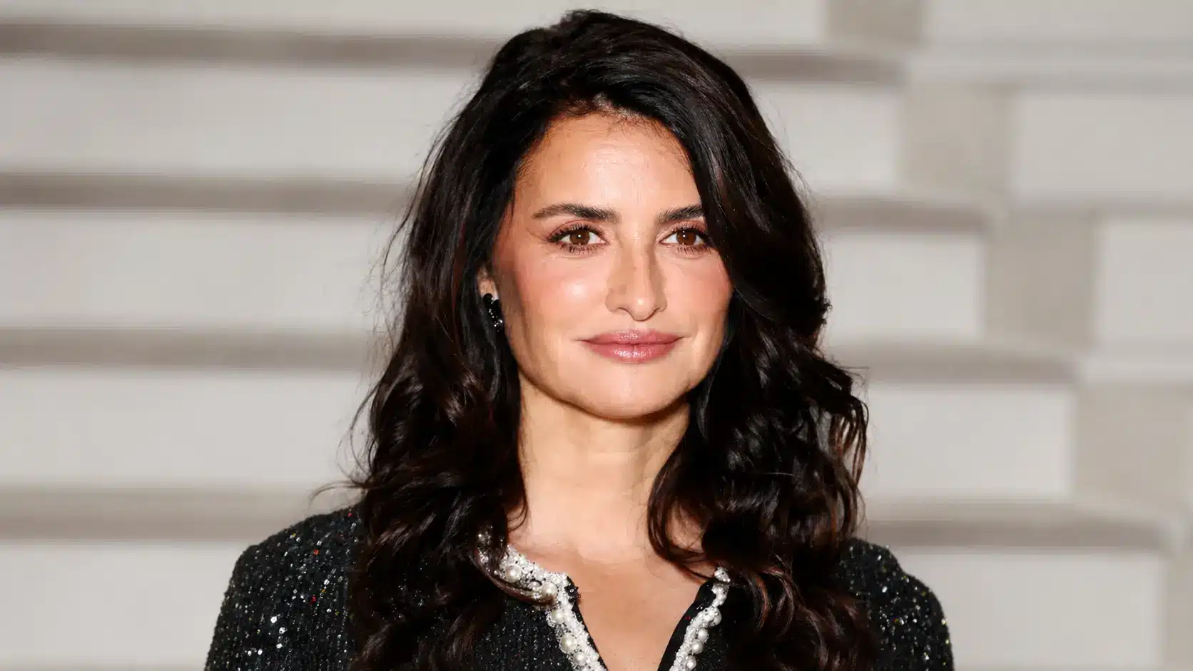 Penélope Cruz with dark wavy hair smiles in a sparkling black outfit, posing against a soft white event backdrop.
