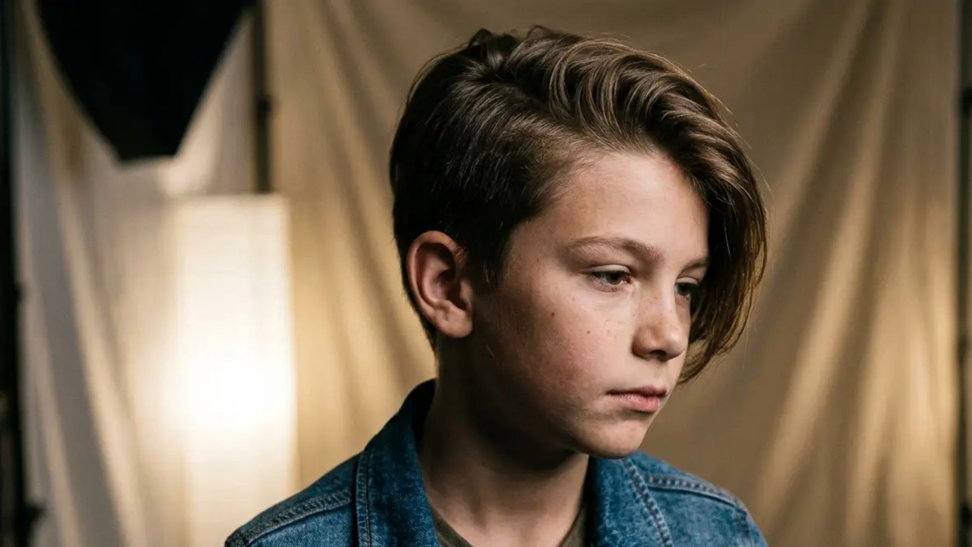 Boy with popstar-inspired layered haircut featuring voluminous side-swept waves, wearing a jacket in a studio-style portrait setting.