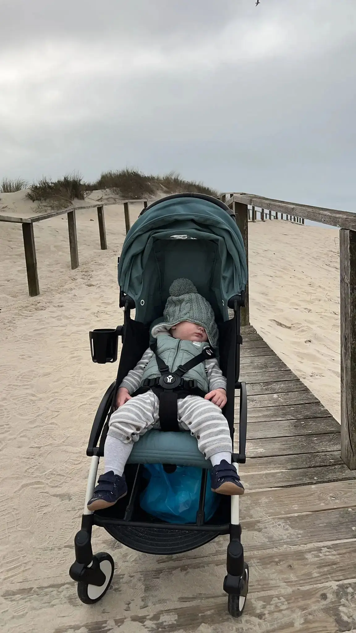 Baby sleeping in stroller on wooden boardwalk at sandy beach under cloudy sky