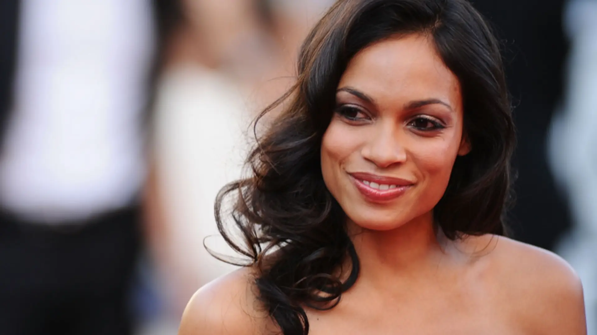 Rosario Dawson wearing a red strapless dress with wavy dark hair posing at a formal event with a blurred background