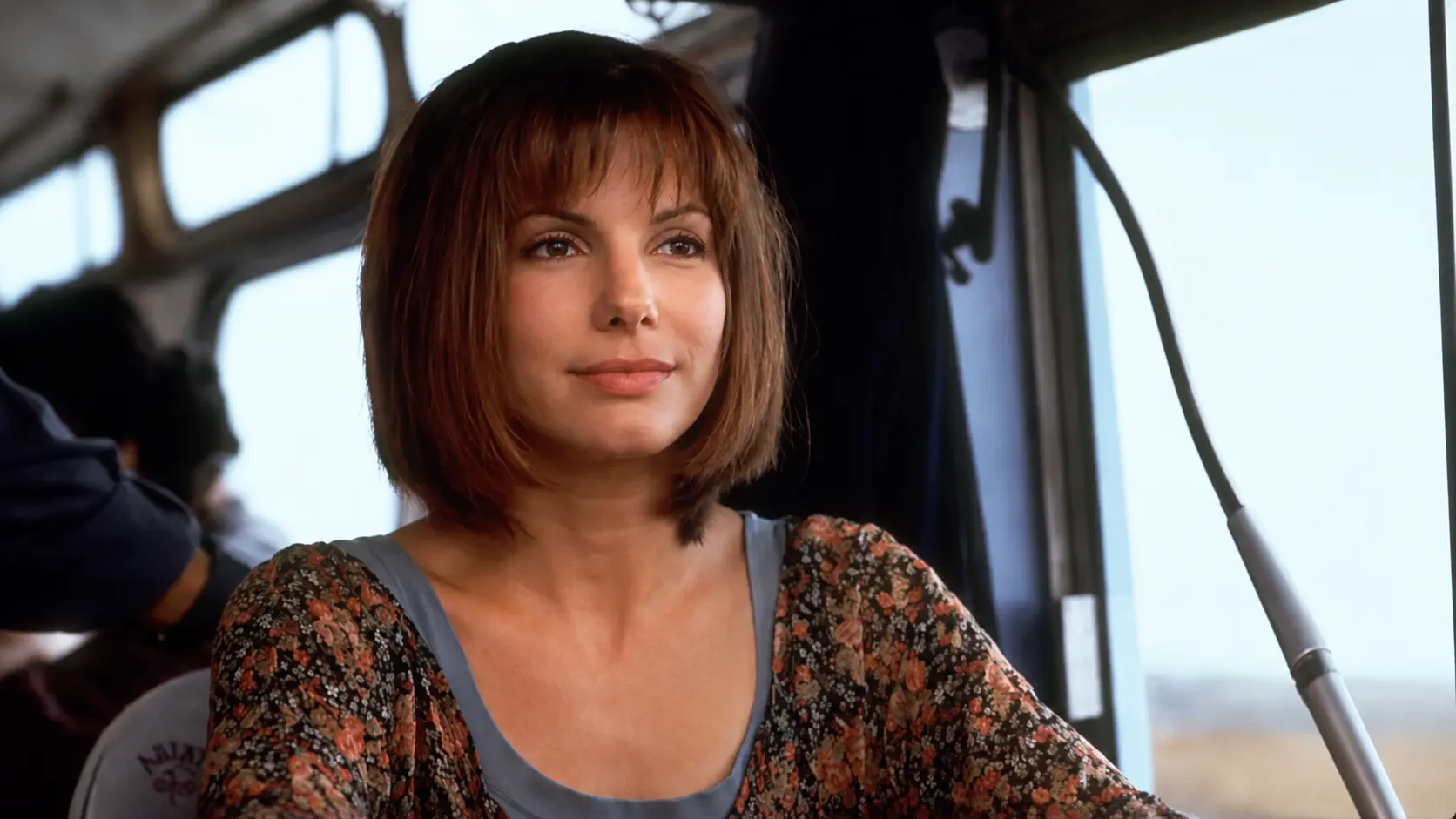Sandra Bullock with short brown hair sitting on a bus, wearing a floral top, with soft natural light coming through the window.