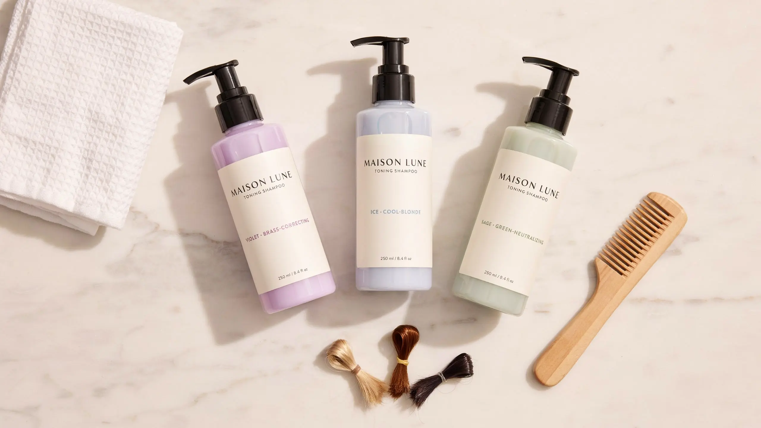 Toning shampoo bottles with blonde hair samples, towel, and wooden comb arranged on a marble surface for hair care.