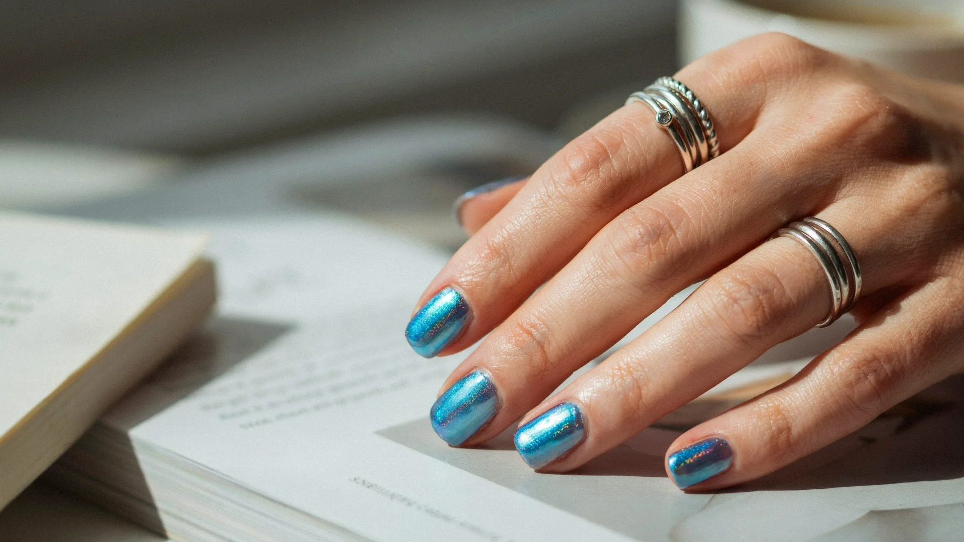 Stylish aqua blue chrome nails featuring a glossy reflective texture on a hand placed over a book surface
