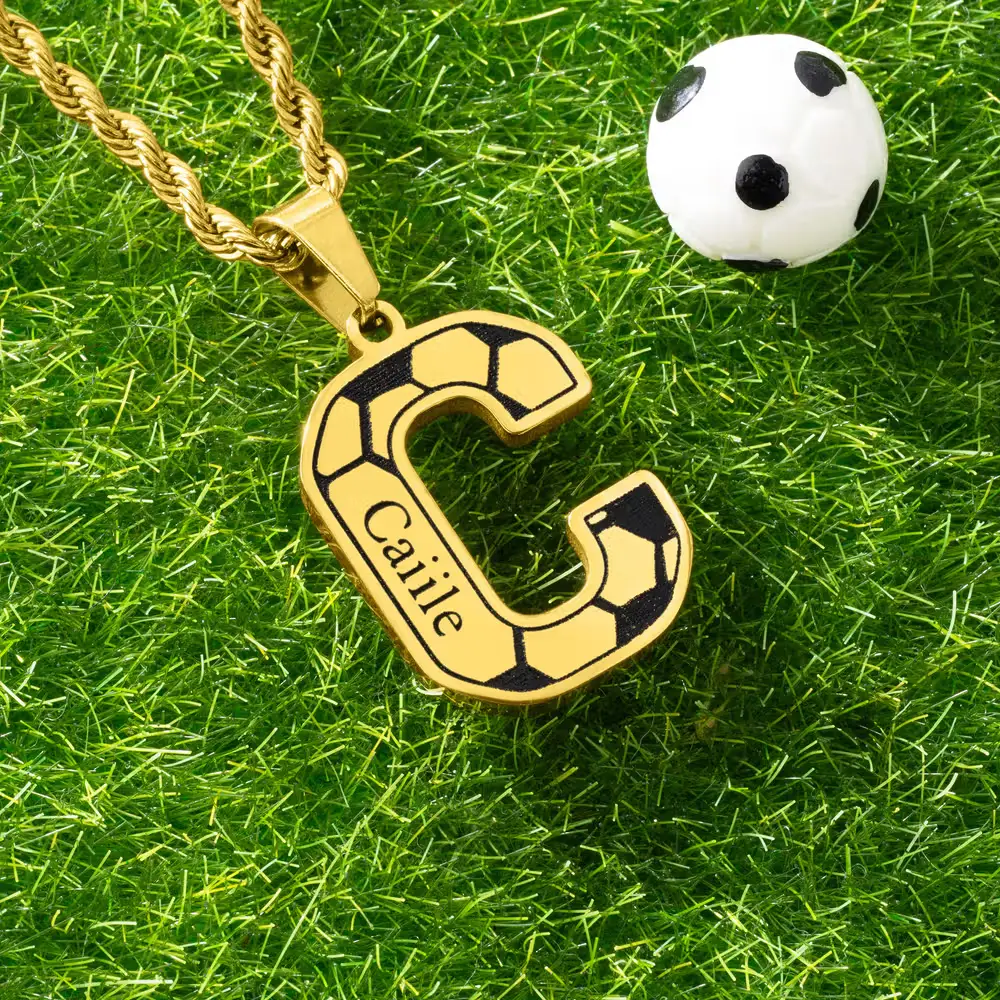Gold chain with soccer-inspired letter C pendant on grass next to small soccer ball