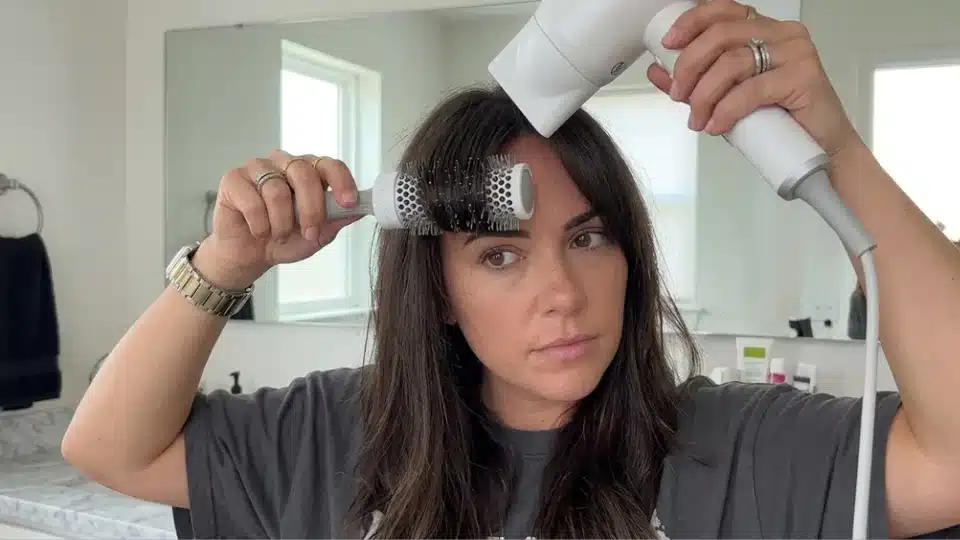 Woman styling curtain bangs at home with a round brush and blow dryer creating a soft outward curve with natural volume