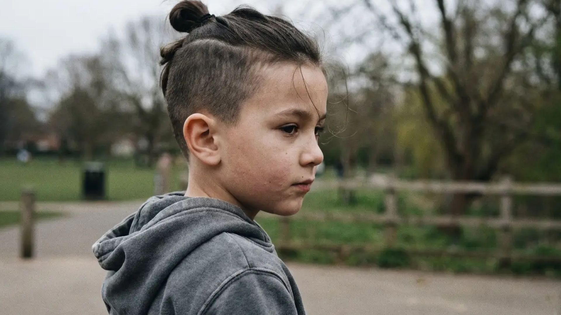 Boy with top knot hairstyle and clean fade on sides, shown in side profile outdoors with blurred background.