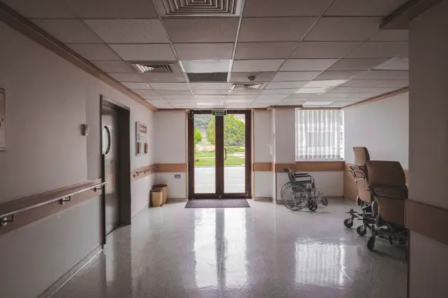 Empty hospital corridor with wheelchair and office chairs near large window, natural light through glass doors