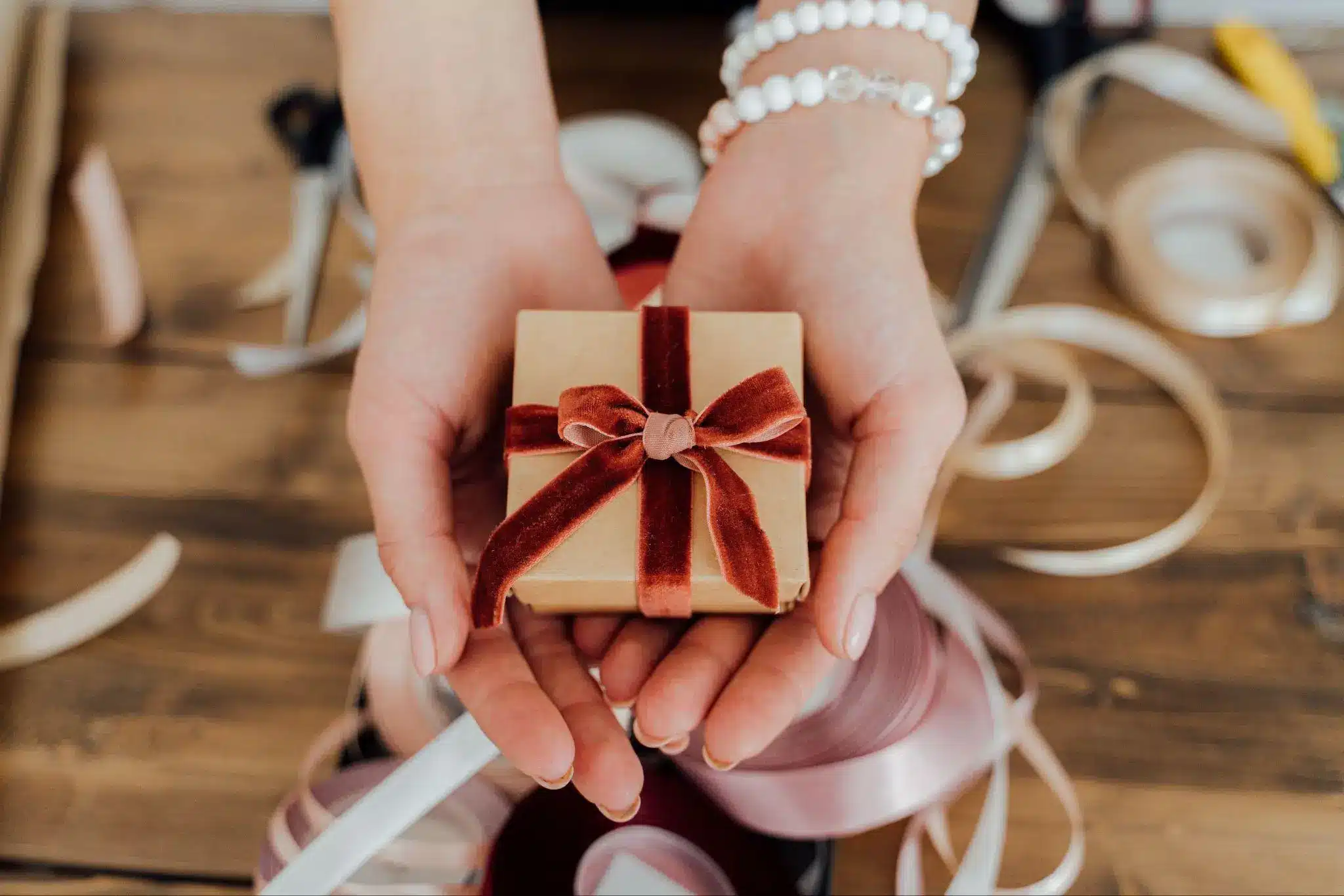 Hands holding small gift box with red velvet ribbon surrounded by ribbons on wooden table